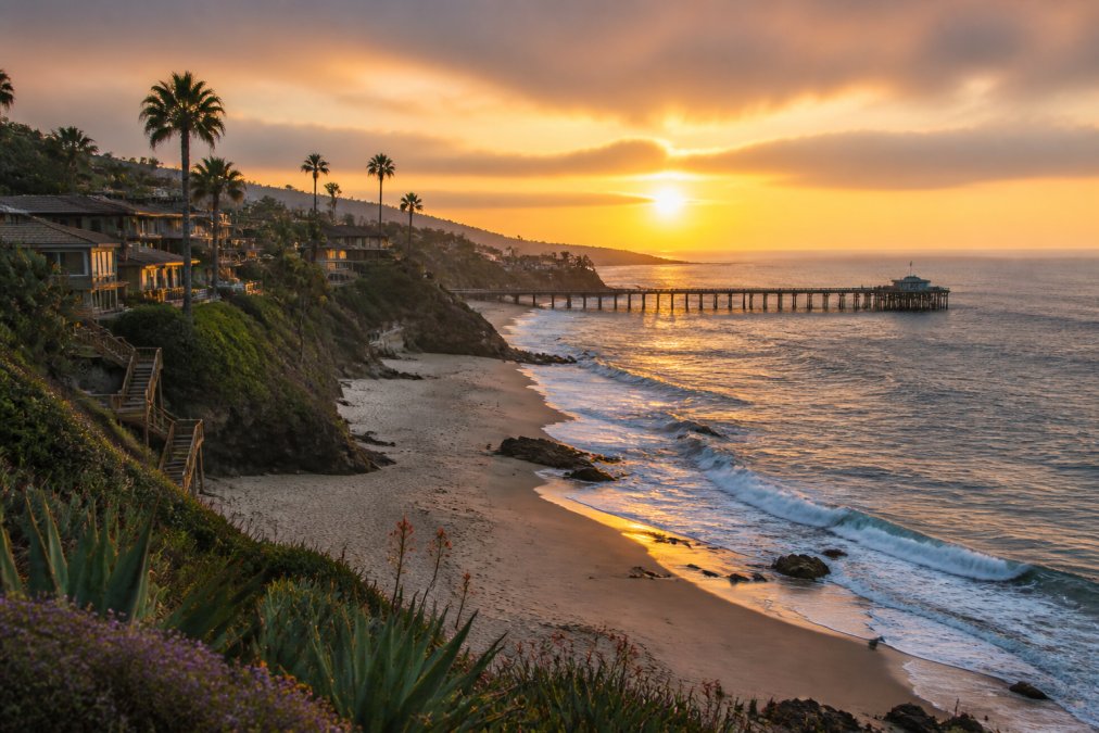 Panoramic sunset view of a Southern California beach town with golden-hour light illuminating sandy shores, colorful beach houses perched on cliffs, and surfers riding final waves of the day