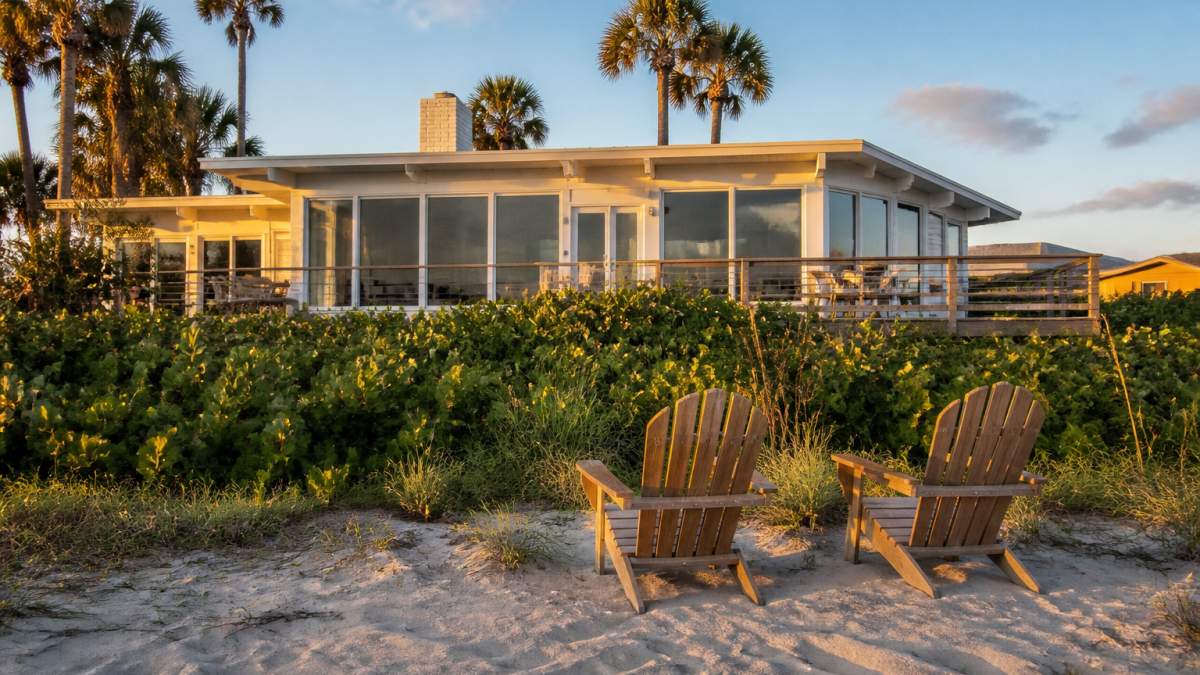 A restored 1950s beachfront home viewed from the sand at golden hour, with wooden deck chairs facing the ocean and sea grapes framing the dune line