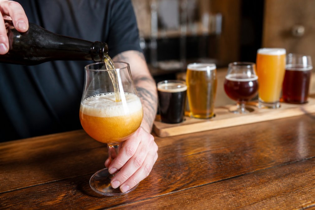 A close-up photo of a bartender pouring a craft beer into a tulip glass on a wooden taproom bar, with a flight of different beer glasses in the background showing varied shapes