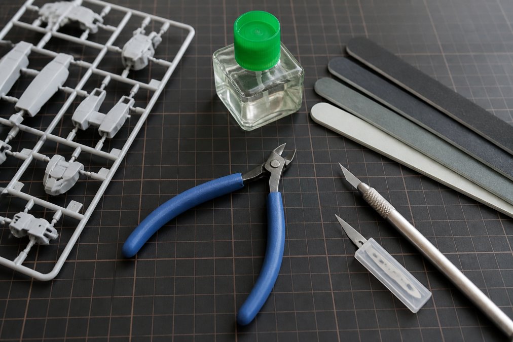 Flat lay of essential Gunpla building tools arranged neatly on a clean workspace: precision nippers, hobby knife with extra blades, assorted sanding sticks in graduated grits, small container of plastic cement, and a sprue from an assembled model kit