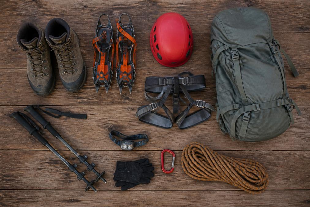 Flat lay of organized mountaineering gear on a worn wooden floor including stiff boots, crampons, a red climbing helmet, harness, trekking poles, a headlamp, and a packed daypack, photographed from directly above in natural daylight