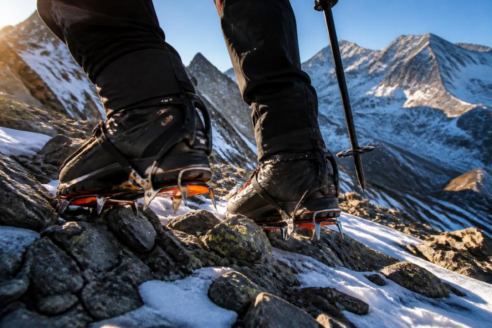 Close-up of a climber's feet wearing rigid mountaineering boots with C2 crampons attached, standing on a steep rocky alpine trail with snow patches, shot in bright morning alpine light