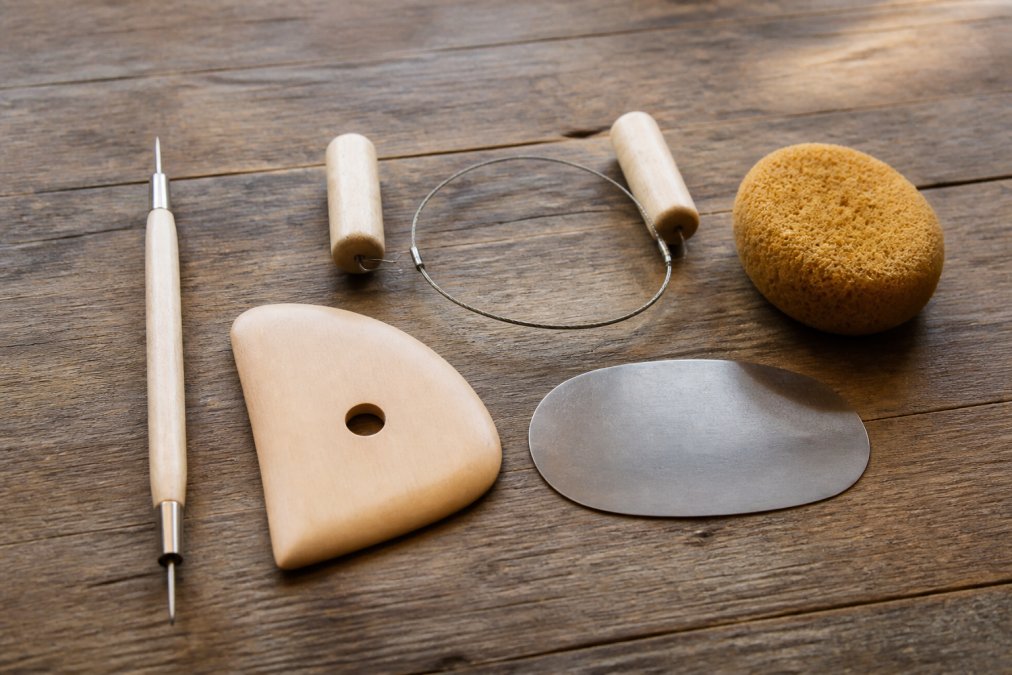 A flat lay of essential pottery tools arranged on a rustic wooden table: a needle tool, a wooden rib, a wire cutter with wooden toggles, a metal scraper, and a natural sponge, all bathed in soft window light