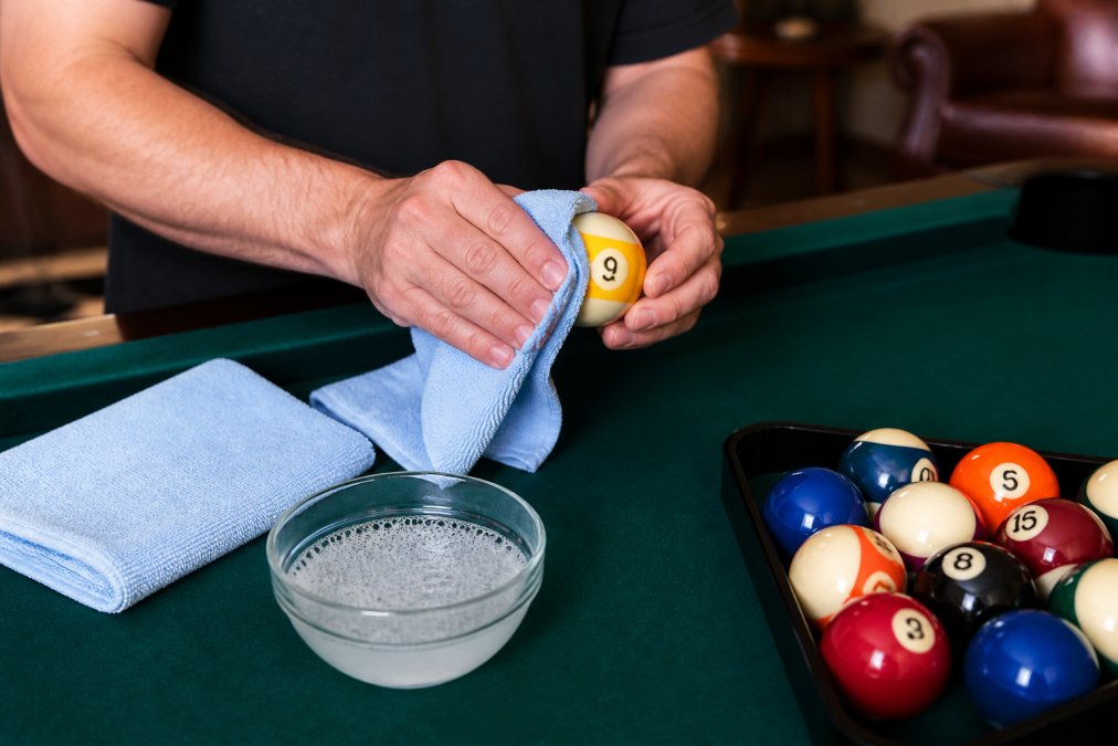 A person's hands gently wiping a billiard ball with a soft microfiber cloth over a green felt table, with a small bowl of mild soapy water and a towel nearby in a home game room