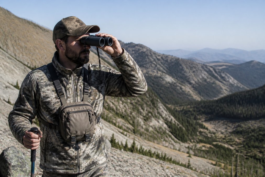Hunter in mountainous backcountry terrain wearing chest-mounted binocular harness, binoculars raised to eyes, demonstrating proper weight distribution and one-handed access during long-range glassing session with distant ridgelines visible