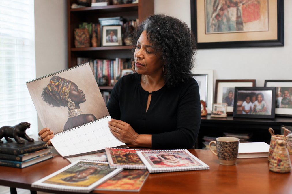Person thoughtfully selecting and reviewing a Black history calendar in a warm home office setting, surrounded by cultural artwork, heritage books, and framed photographs of family, natural window lighting