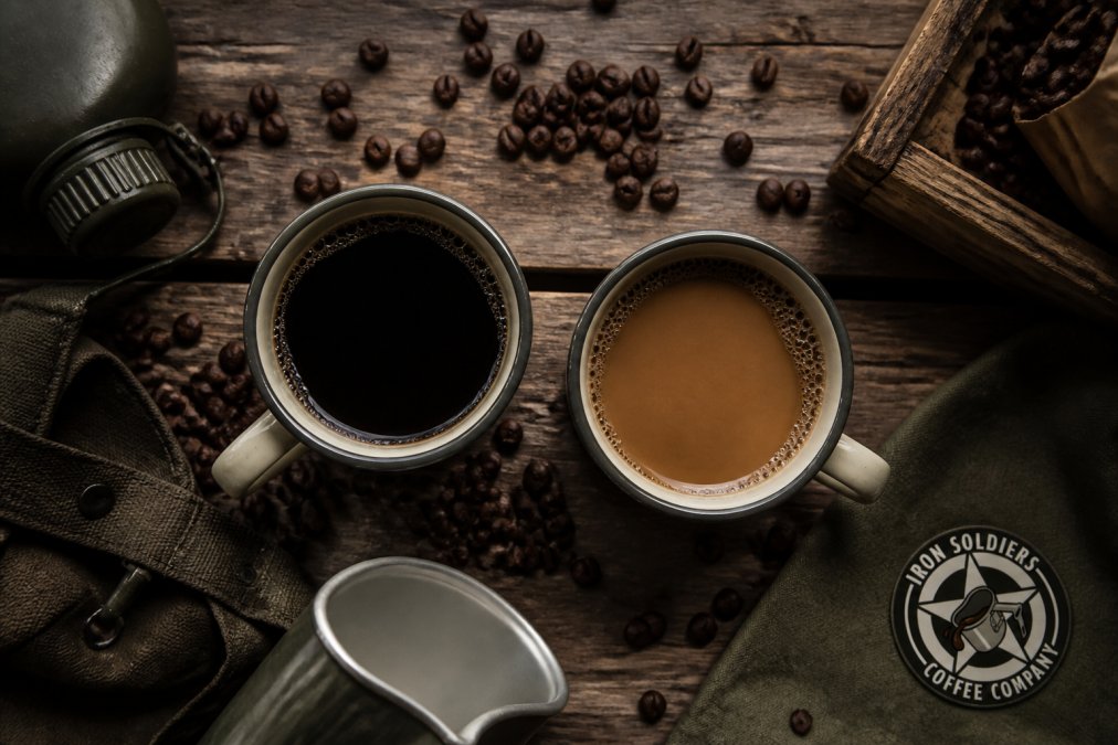 Overhead shot of two vintage-style coffee mugs on a weathered wooden table, one filled with dark roast coffee and one with medium roast, surrounded by scattered whole coffee beans and a military-style canteen cup