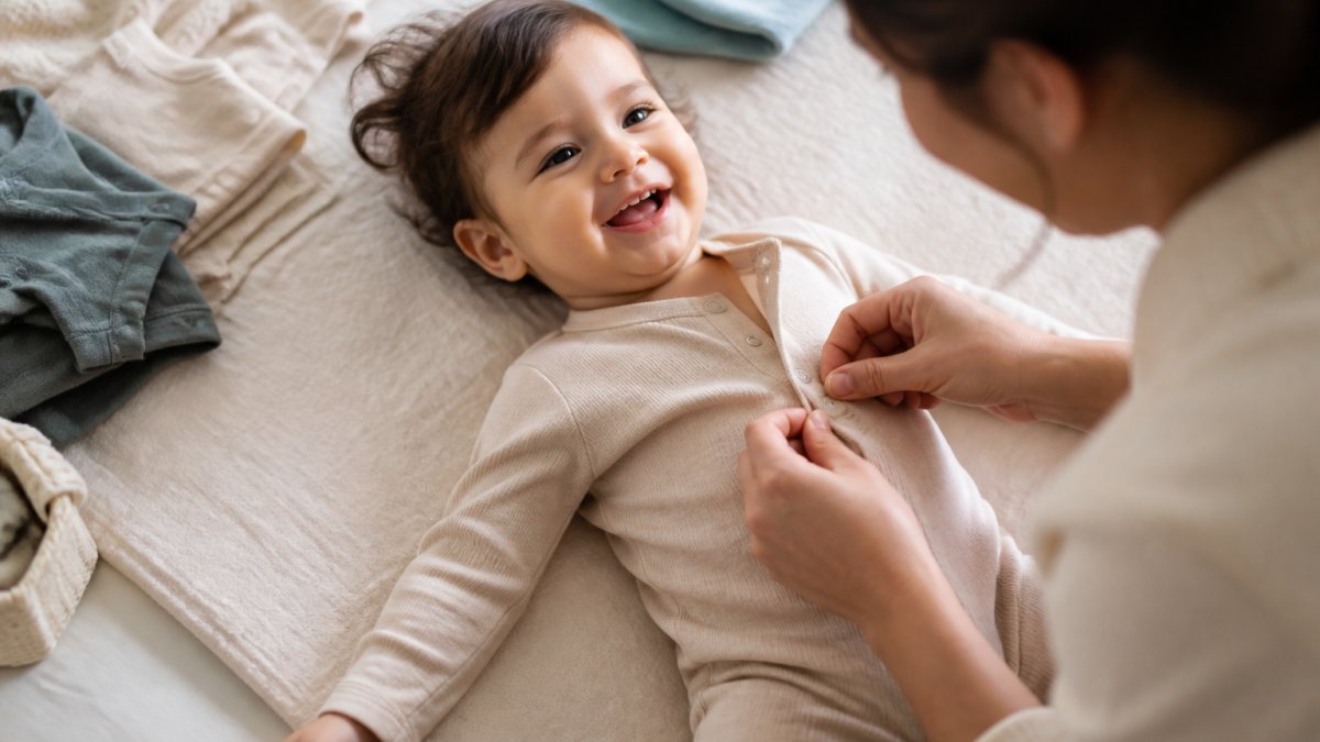 Parent dressing a smiling toddler in soft cotton clothing, showing the fabric texture and ease of putting on a romper with snap closures in natural home lighting