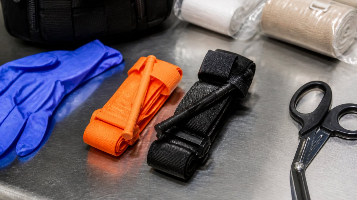 Orange and black tactical tourniquets laid out on a stainless steel bench with nitrile gloves and trauma shears