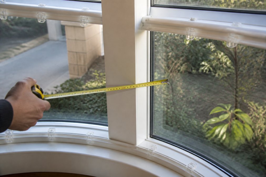 Measuring tape held against a bay window frame showing the angle and projection depth, with natural daylight illuminating the window recess and the tape clearly visible against the frame