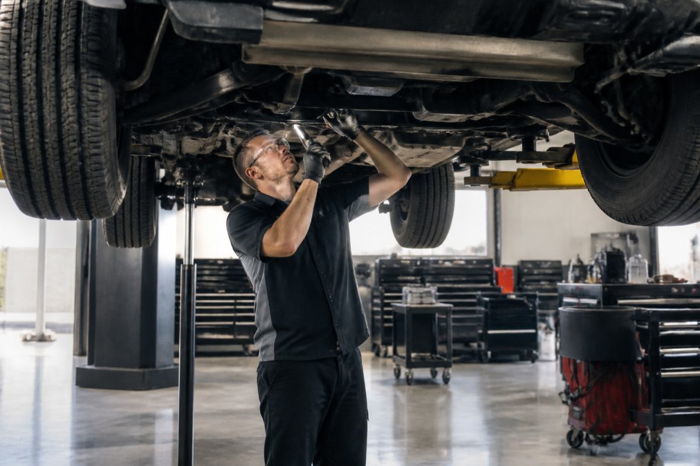 An ASE-trained technician in a clean, well-lit auto repair shop bay carefully inspecting the undercarriage of a raised truck on a hydraulic lift, with organized tool chests and diagnostic equipment visible in the background