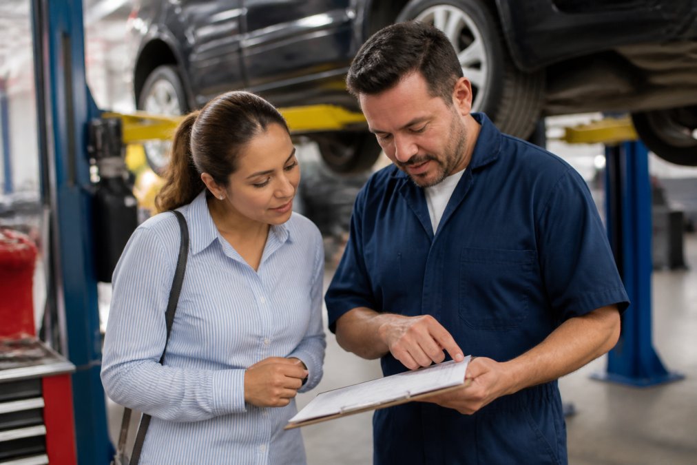 A customer and a mechanic in a bright, organized auto repair shop reviewing a printed repair estimate on a clipboard together, both engaged in conversation with the vehicle visible on the lift in the background