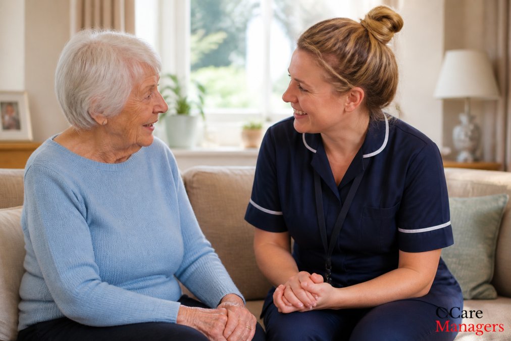 A professional female carer having a warm, respectful conversation with an elderly woman in a bright, comfortable living room with soft natural light