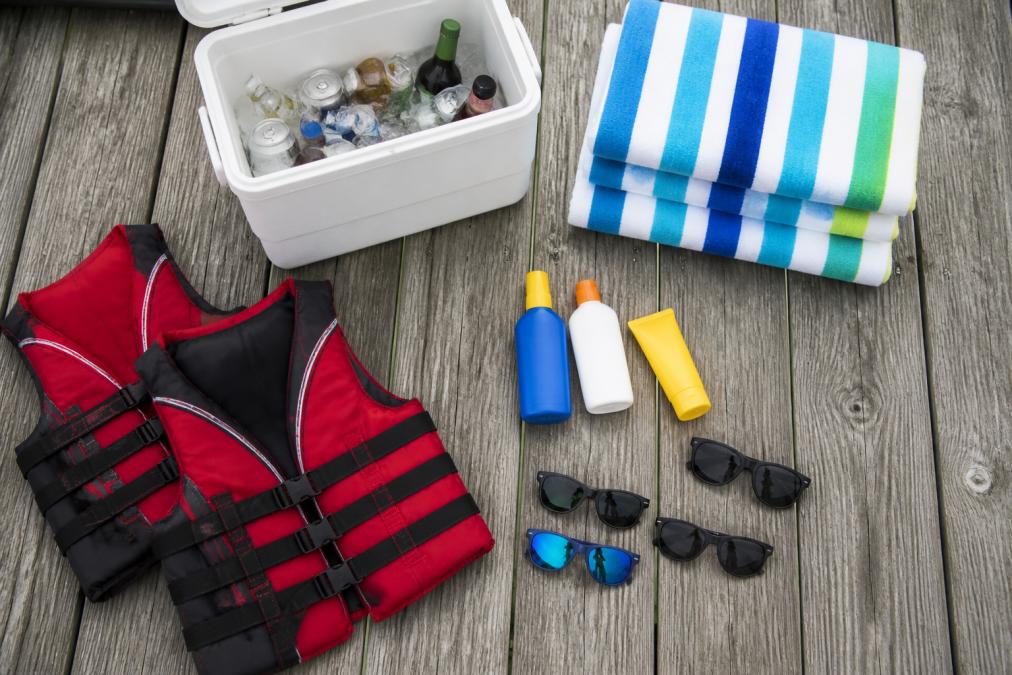 Overhead shot of essentials laid out on a dock: sunscreen bottles, life jackets, cooler with beverages, colorful towels, and sunglasses arranged neatly on weathered wood