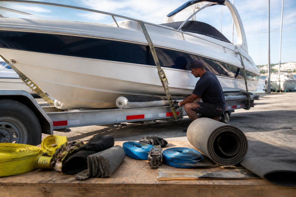 Boat owner securing vessel with heavy straps and foam cradle supports in marina setting, checking hull integrity and fastening points in natural daylight
