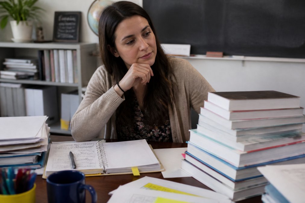 A young teacher sitting at a desk covered with books and lesson plans, looking thoughtfully at a stack of professional development books in a classroom setting