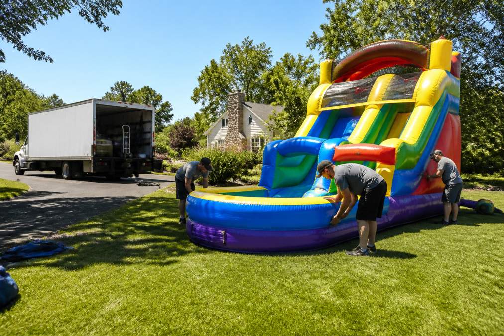 Professional rental team setting up a colorful bounce house and water slide combo at a backyard event venue, with delivery truck visible in background, sunny day with green grass and clear sky