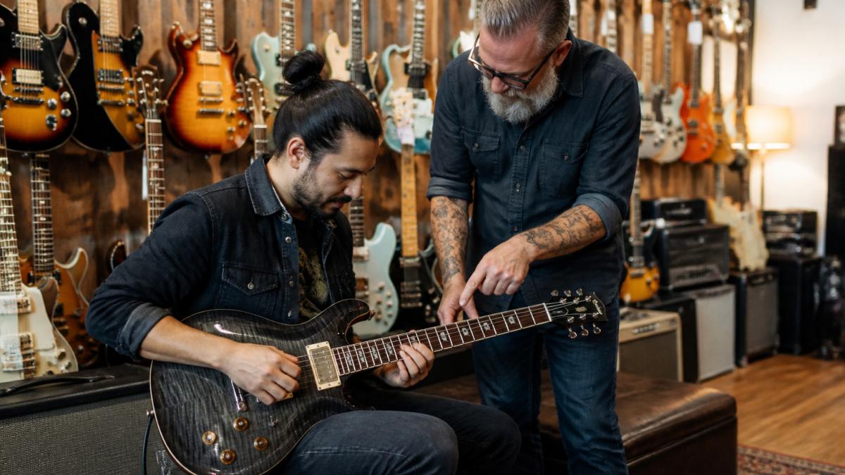 A musician seated in a warm, well-lit independent guitar shop, holding a boutique electric guitar across their lap while a staff member points to the setup, with rows of handcrafted guitars hanging on the wall behind them