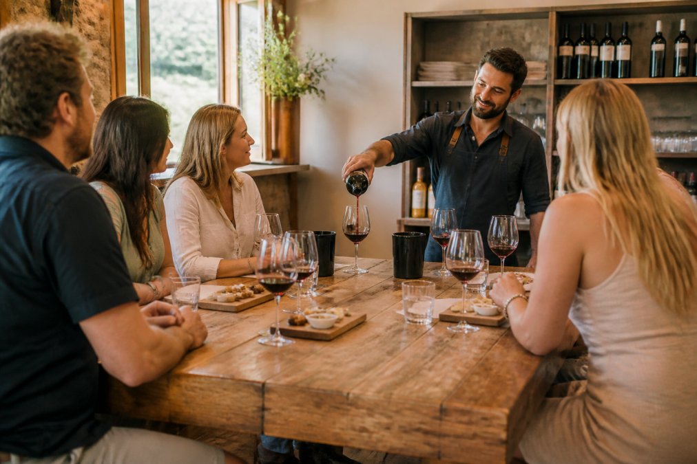 A warm, inviting photo of a small group of friends tasting wine at a rustic wooden cellar door counter, with a winemaker pouring a glass of red wine in the background. Natural light streams in from a nearby window, highlighting the bottles on the shelf.