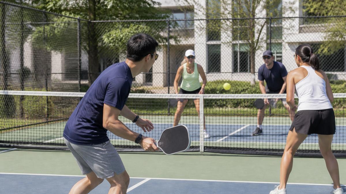 Diverse group of office employees playing pickleball on an outdoor court during a company team-building event, with branded paddles visible in action and natural sunlight illuminating the court