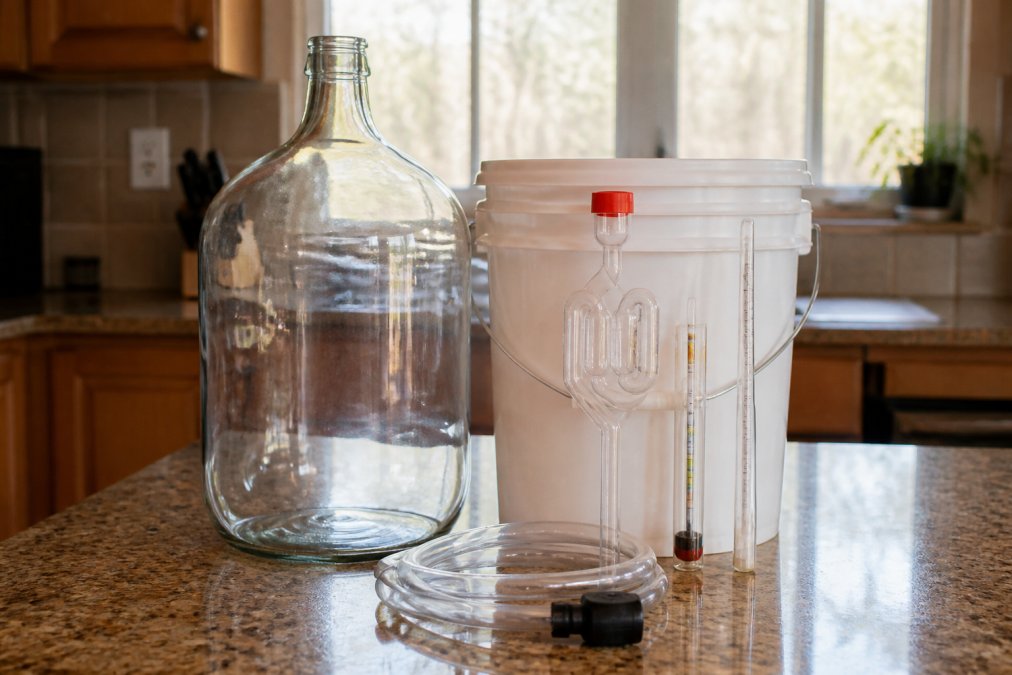A home brewer's kitchen counter with a glass carboy, airlock, siphon, hydrometer, and sanitizer bucket arranged neatly, natural window light