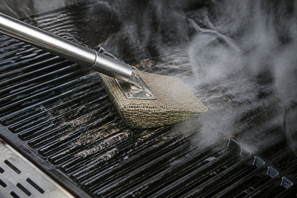 Close-up of steam rising from grill grates as a bristle-free Kevlar scrubber removes charred residue, showing the cleaning action with visible steam and grime lifting