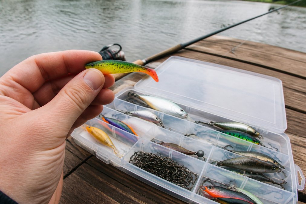 A close-up shot of a hand holding a small, colourful soft plastic fishing lure next to a tackle box filled with various hard lures and hooks, set on a wooden dock with a blurred river background