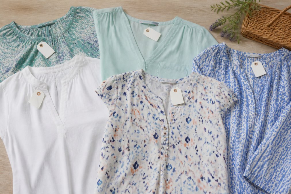 A neatly arranged flat lay of affordable women's blouses and tops in soft mint, white, and blue tones on a light wooden surface, with small price tags visible against the fabric