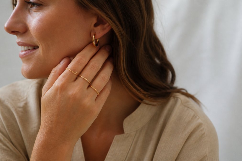Close-up of a woman's hands wearing delicate hoop earrings and stacked rings, styled with neutral tones and natural lighting