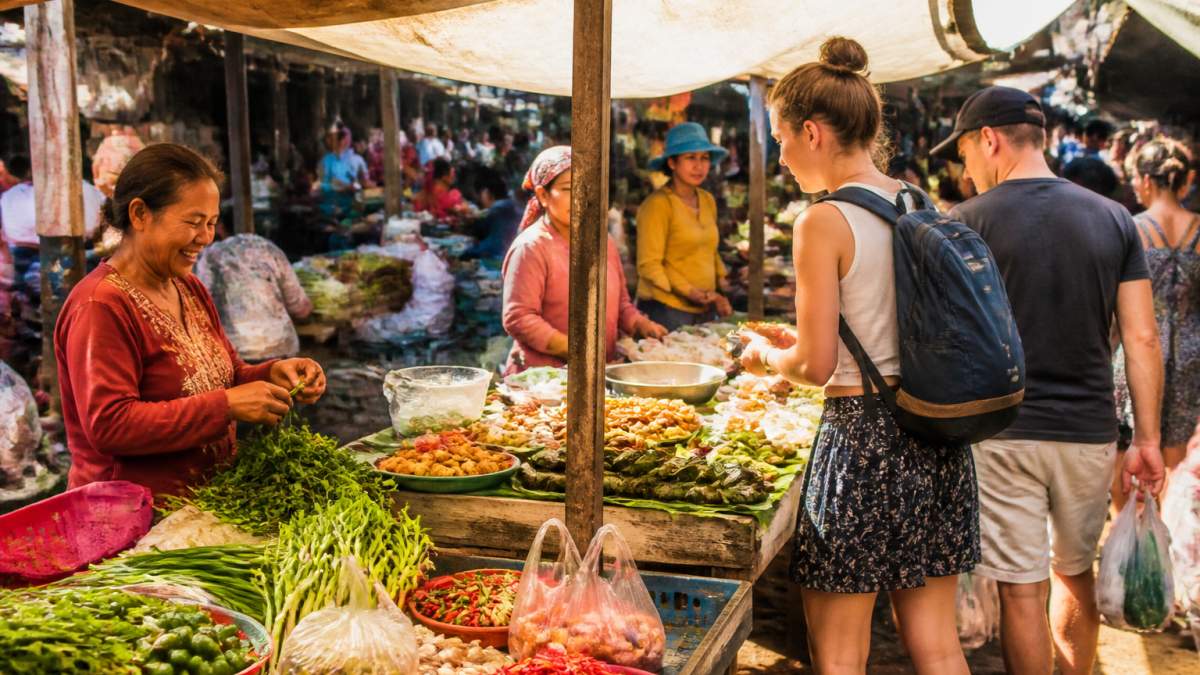 Vibrant street market scene with local vendors selling colourful produce, fresh herbs, and street food in a busy Southeast Asian setting, with tourists and locals browsing wooden stalls under bright sunlight