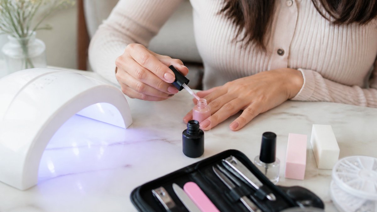 Close-up of a woman's hands applying gel polish from a starter kit at a clean, bright home table with a small LED lamp nearby