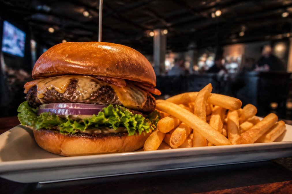 A juicy, hand-crafted burger with a toasted brioche bun and a side of french fries on a plate, with a sports bar background showing big screens and a casual dining atmosphere