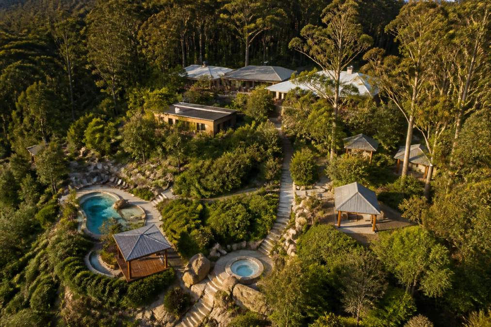 Aerial view of a luxury wellness retreat nestled in Australian bushland with manicured gardens, thermal pools, meditation pavilions, and spa facilities visible among native trees