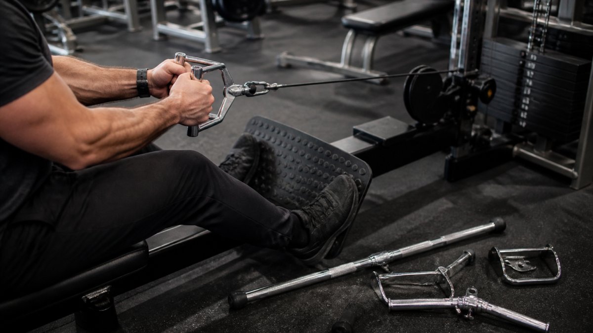 Close-up of a lifter's hands gripping a V-grip cable row handle on a low pulley cable machine in a commercial gym, with other attachments resting on the floor nearby