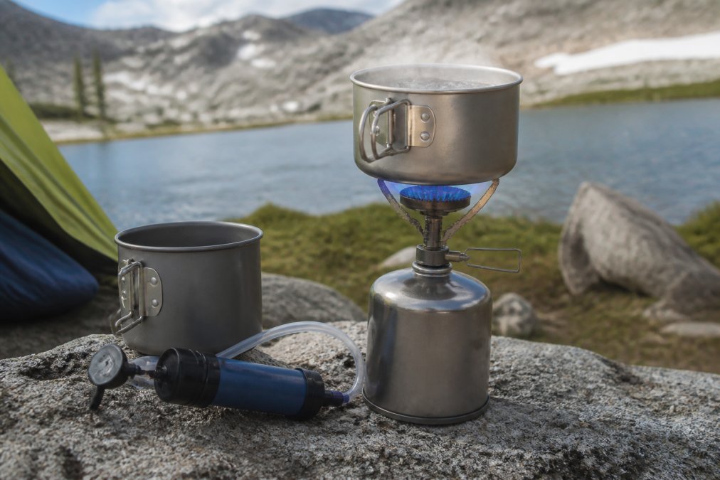 Close-up of a compact silver canister stove with a blue flame boiling water in a titanium pot on a flat granite rock at a backcountry campsite, with a Sawyer Squeeze water filter and a gray cook pot visible beside it in soft morning light