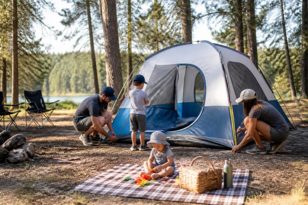 A family of four pitching a large dome tent together at a sunny campsite, with a toddler playing nearby on a picnic blanket