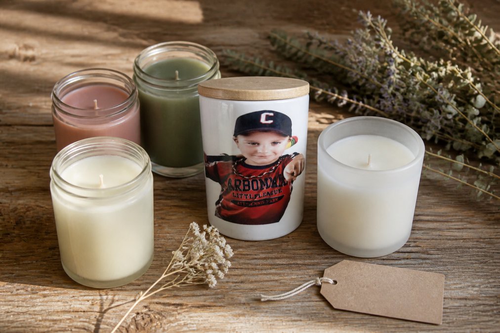 A styled flat-lay of several hand-poured scented jar candles in frosted and clear glass vessels arranged on a worn wooden surface with dried botanicals, a small kraft paper gift tag, and soft natural window light falling across the scene
