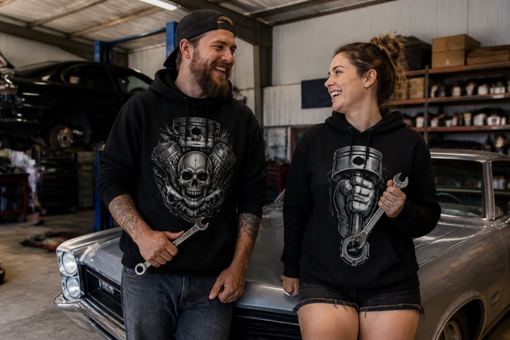 Smiling couple in automotive hoodies holding wrenches by a classic car, representing some of the best car enthusiast gift