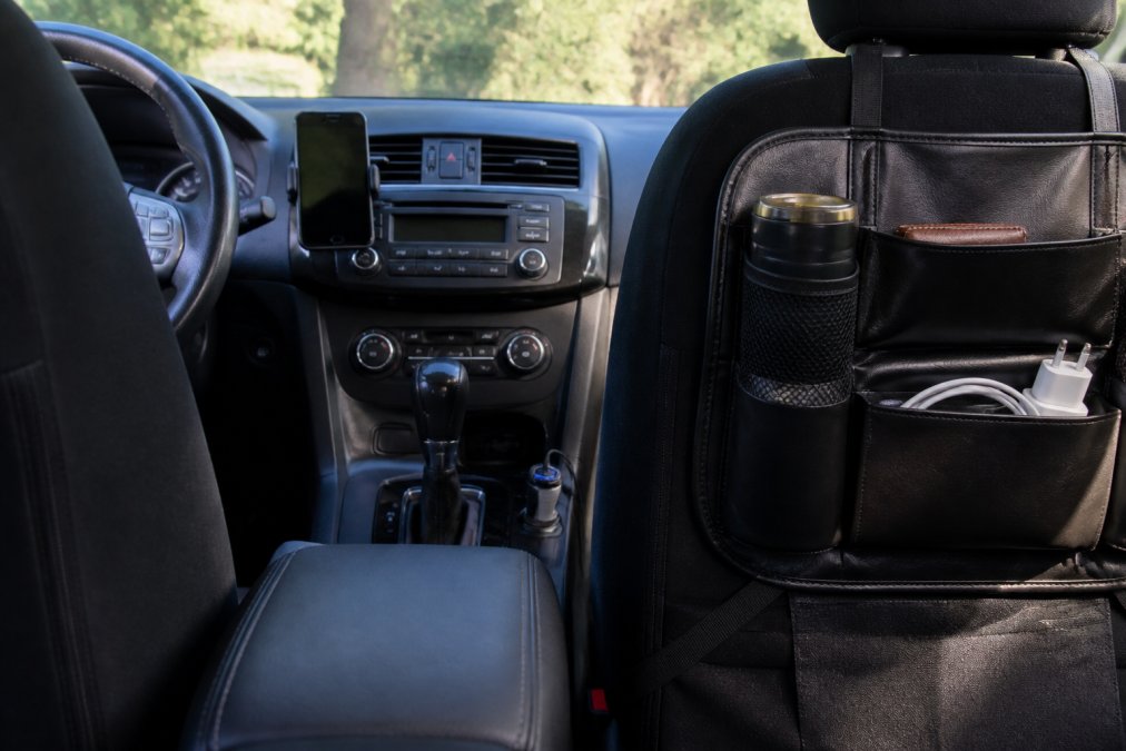 Dashboard of a modern car with various affordable gadgets installed: magnetic phone mount holding a smartphone, FM transmitter in the 12V outlet, and interior organizer visible on the seat back, with natural daylight streaming through windshield