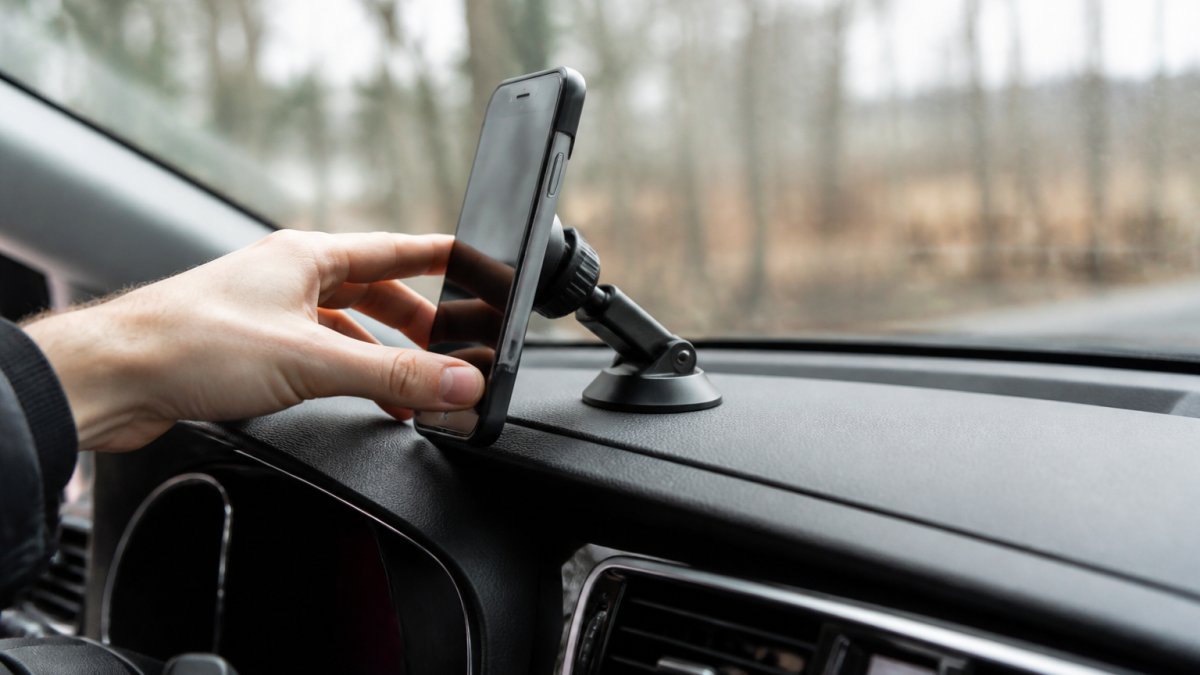 A driver's hand pressing a magnetic phone mount attached to a textured car dashboard, with a smartphone secured in the cradle, photographed in daylight through the windshield