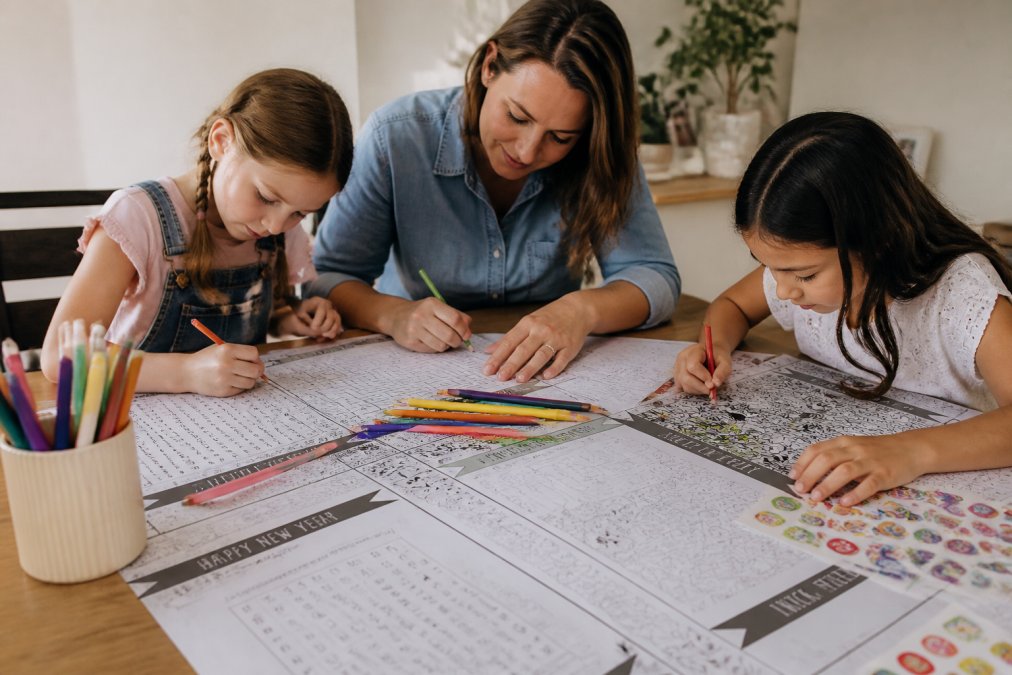 Two children and one adult bent over a table covered with various printable activity materials, word searches, crossword puzzles, sticker sheets, and coloring pages, all working with focused concentration, colored pencils and markers scattered across the table surface, warm indoor lighting