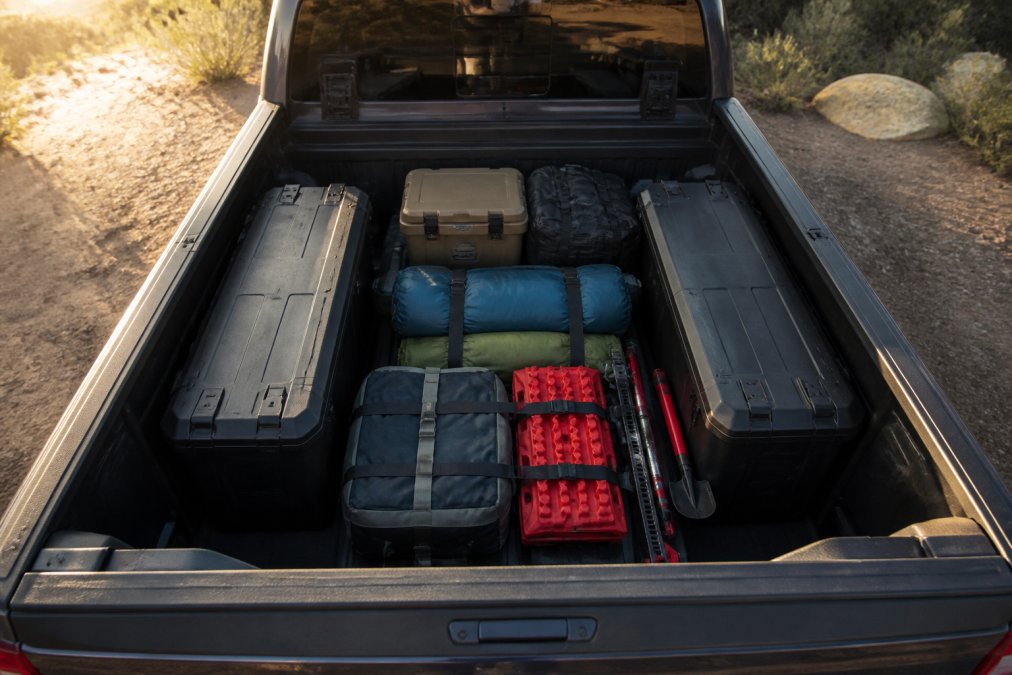 Overhead view of a truck bed with two weatherproof storage boxes neatly arranged alongside camping gear, sleeping bags, and recovery equipment secured with cargo straps, morning sunlight illuminating the organized setup