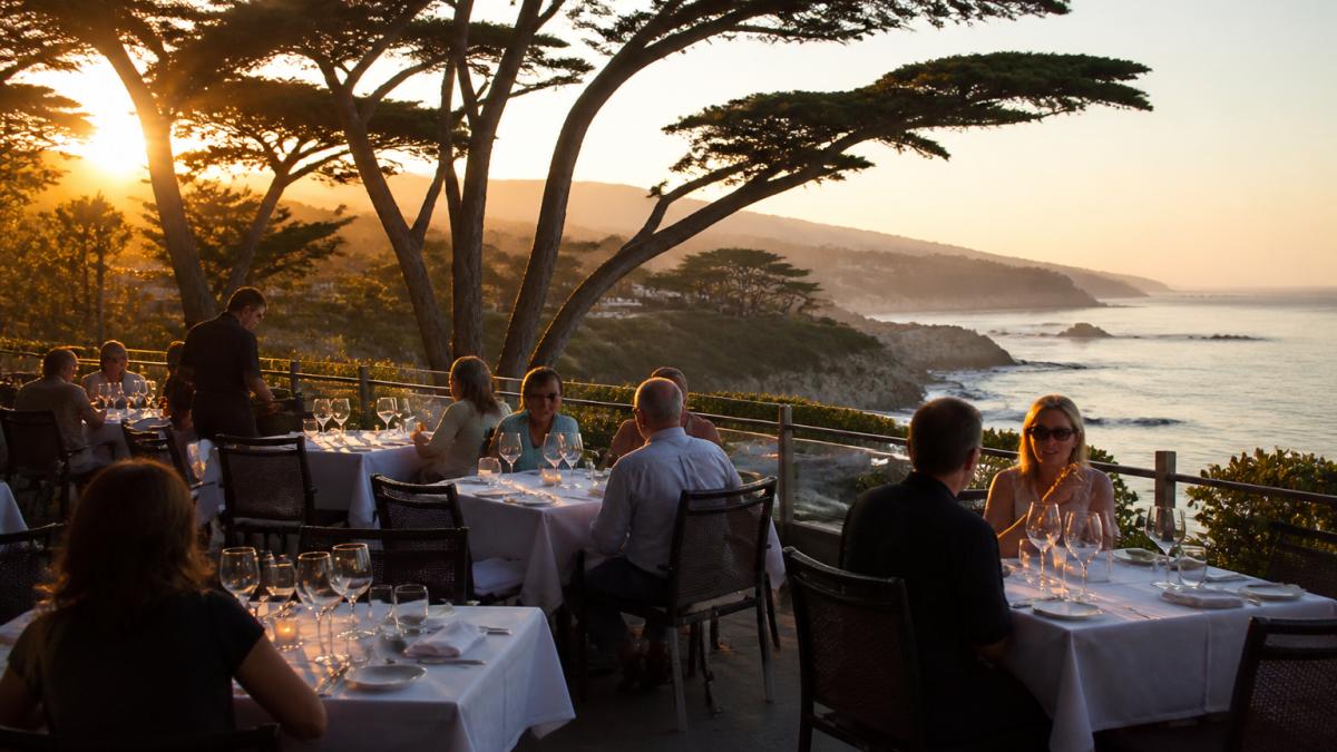 Elegant outdoor patio dining area overlooking the Carmel coastline at golden hour, with candlelit tables, wine glasses catching the light, and diners in conversation against a backdrop of cypress trees and ocean cliffs