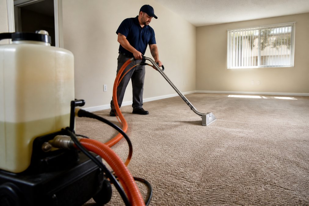 A professional carpet cleaning technician operating a hot water extraction machine on a beige carpet in a vacant rental apartment, with orange hoses connected to a solution tank visible in the foreground and natural daylight coming through a window