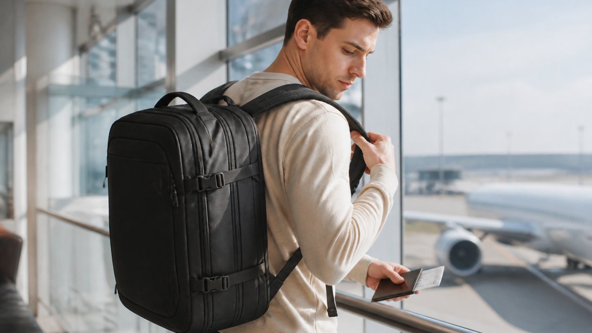 A traveller in a bright airport terminal adjusting the straps of a black carry-on backpack, with a boarding pass in hand and a plane visible through the window behind them