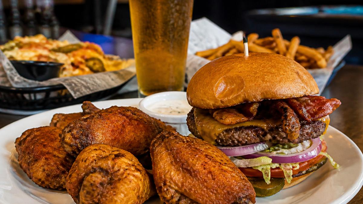 Close-up of crispy golden wings and a loaded burger on a plate with cold beer and appetizers in the background, casual sports bar atmosphere with warm lighting