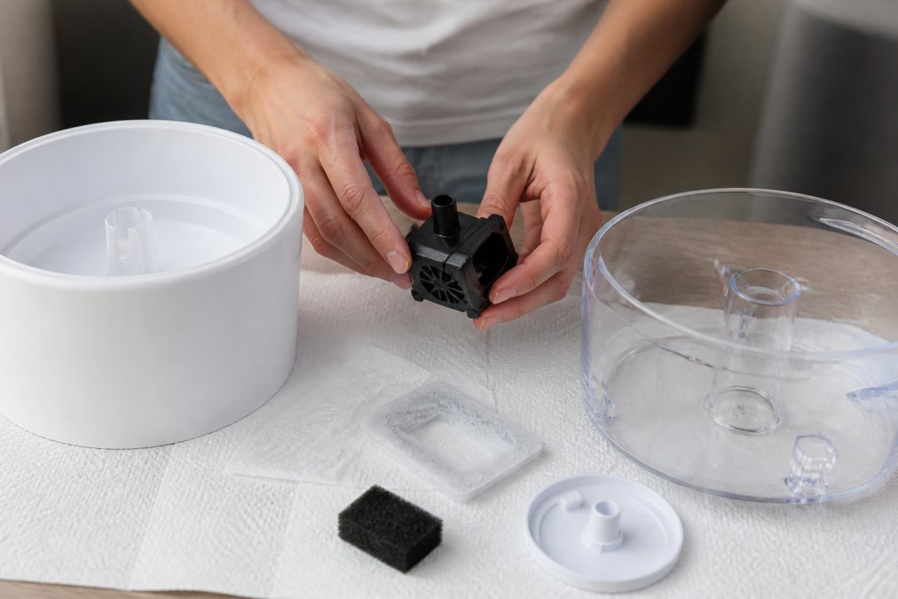 Close-up of hands disassembling a cat water fountain, showing the pump, filter, and reservoir components laid out on a clean surface with paper towels