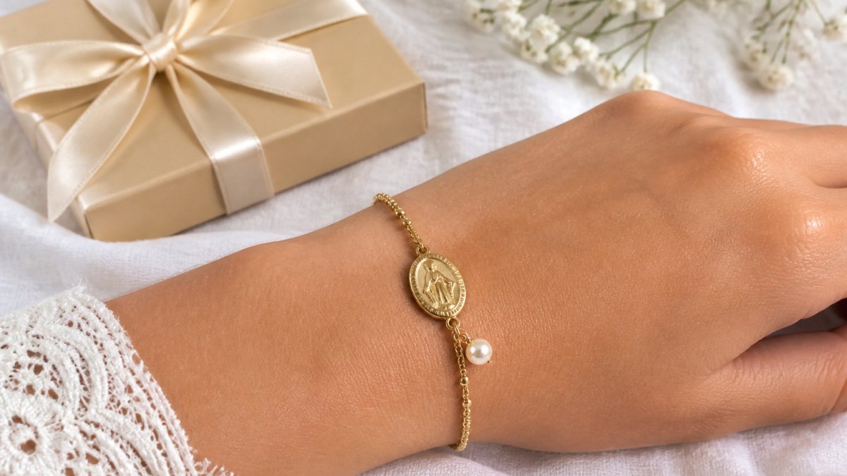 Close-up of a teenage girl's wrist wearing a delicate gold-plated Miraculous Medal bracelet with a small pearl accent, resting on a white linen surface beside a gift box