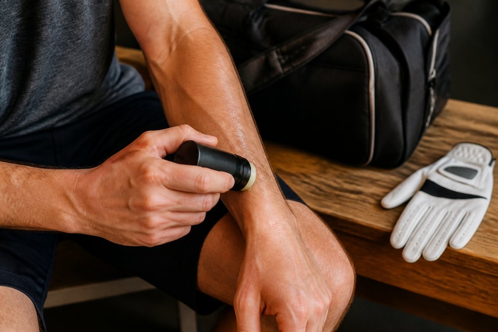 Close-up of an athlete's hands applying a CBD salve stick to a forearm after a workout, with a gym bag and golf glove visible on a wooden bench in the background, warm natural lighting