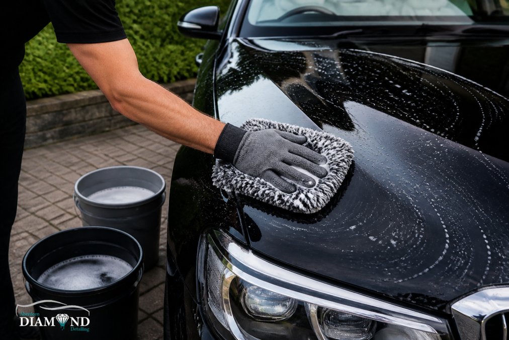 Close-up of a person wearing microfibre gloves hand-washing the glossy bonnet of a dark prestige car with a plush wash mitt and two buckets of soapy water on a driveway